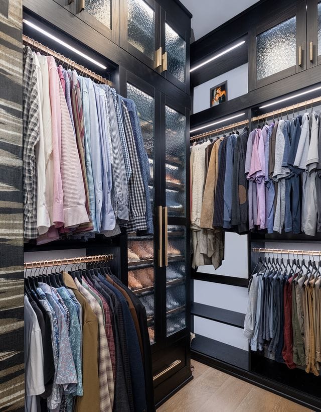Walk in closet designed for his side in dark wood grain finish with custom shoe storage behind cabinet doors with glass inserts created by California Closets